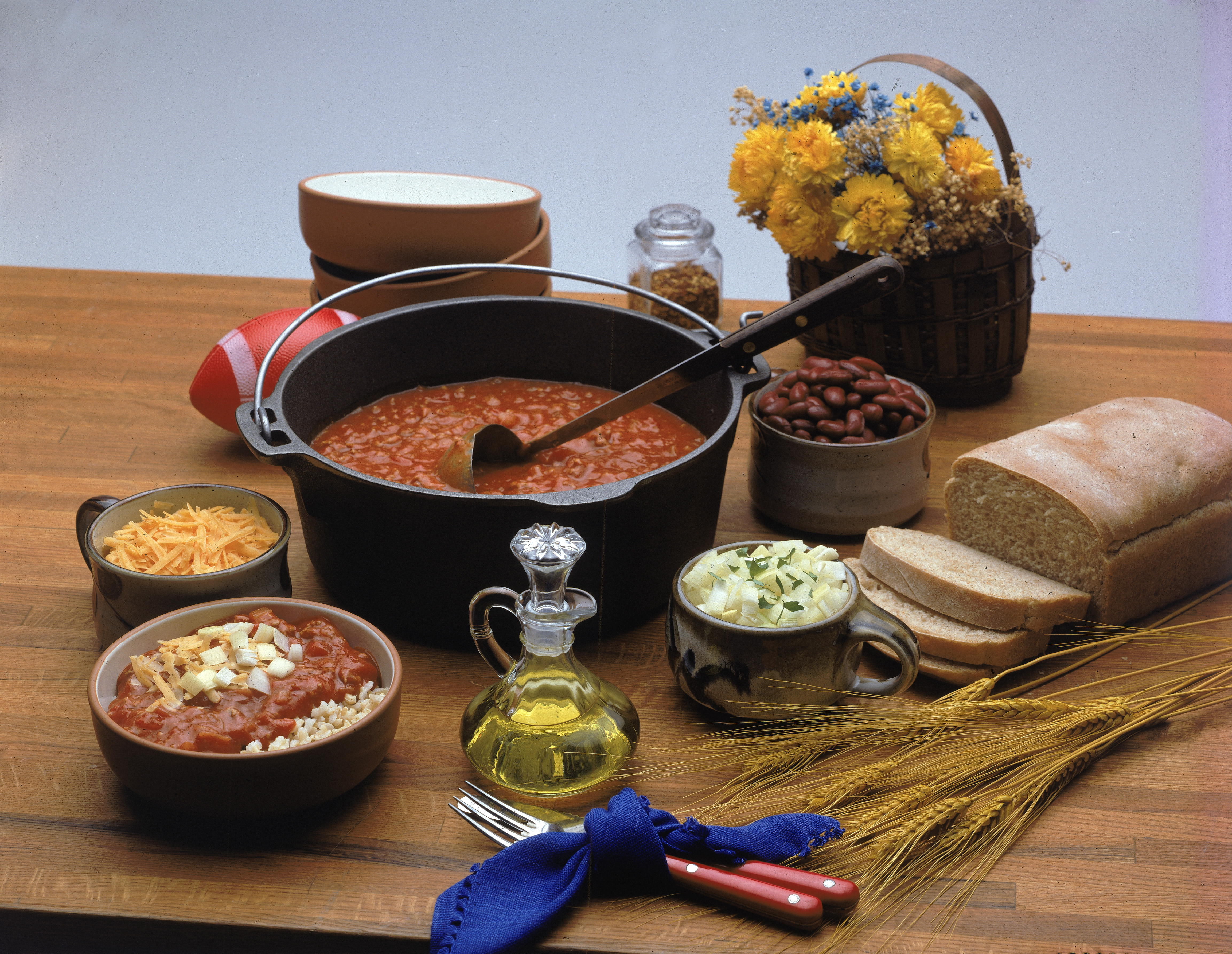 All the items necessary for a Mexican-themed chili feast sit on a wooden surface including a cauldron of chili (with ladle), a loaf of bread, a bowl of brown rice and chili, a bowl of chopped onions, a bowl of shredded cheese, a bottle of oil, a toy plastic football, a basket of dried flowers, a decorative sheaf of wheat, and some utensils and dishes, 1970s. (Photo by Hulton Archive/Getty Images)