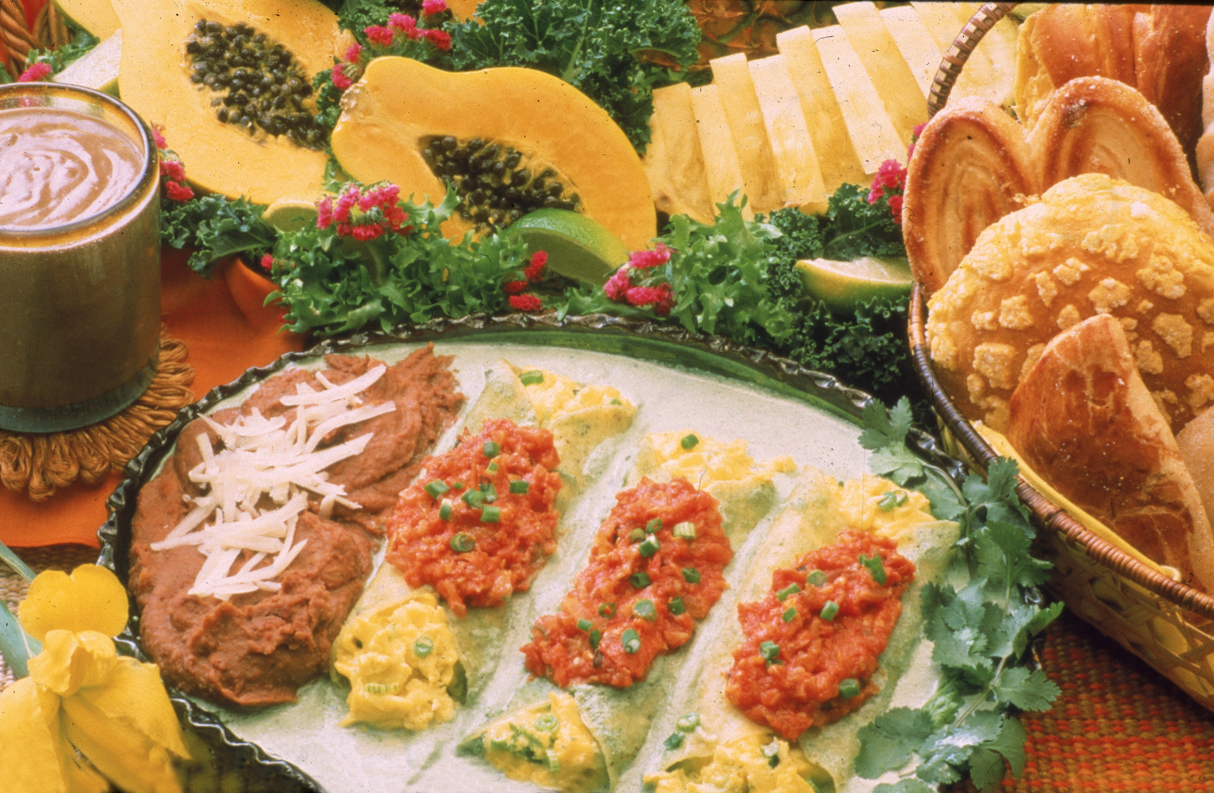 Photograph shows display of Mexican-themed food items including eggs wrapped in tortillas and smothered in salsa, refried beans, breads and pastries, papaya, and lime, 1970s. (Photo by Hulton Archive/Getty Images)