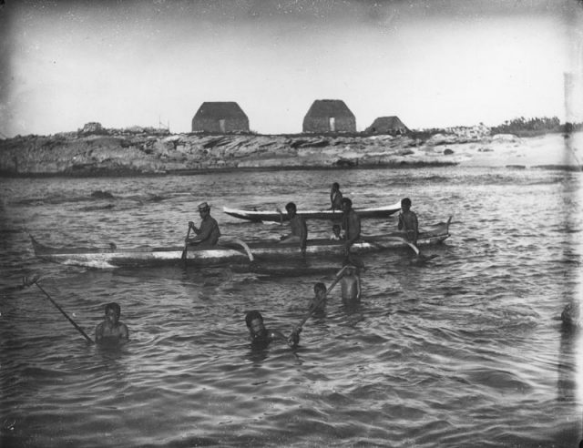 Men and boys fish in a bay, some in outrigger canoes, others in the water with snared fish. Beyond is a rocky shore with three thatched houses. This photo was taken by Francis Sinclair in 1885.