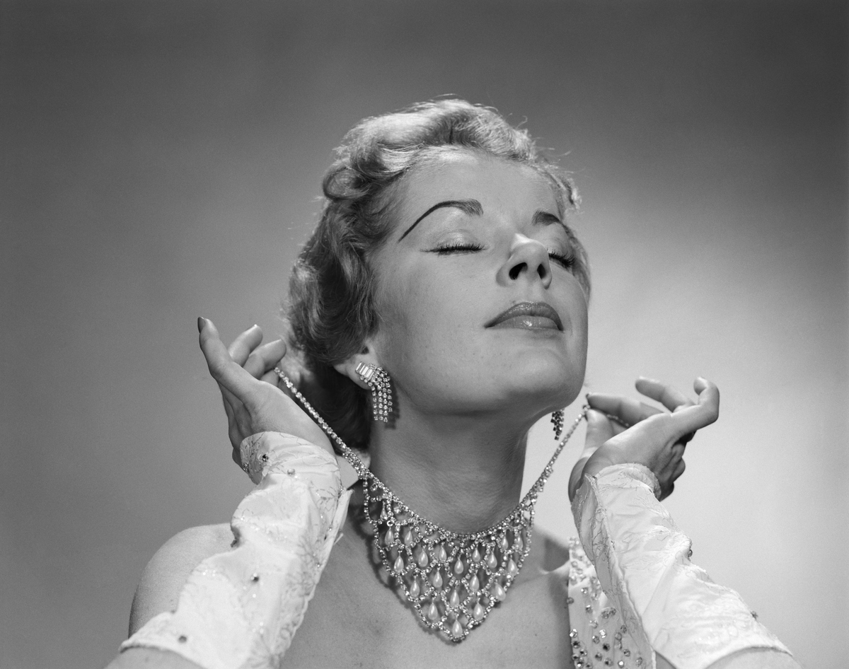 1950s GLAMOROUS WOMAN EYES CLOSED CHIN LIFTED PUTTING ON ELABORATE DIAMOND RHINESTONE NECKLACE 