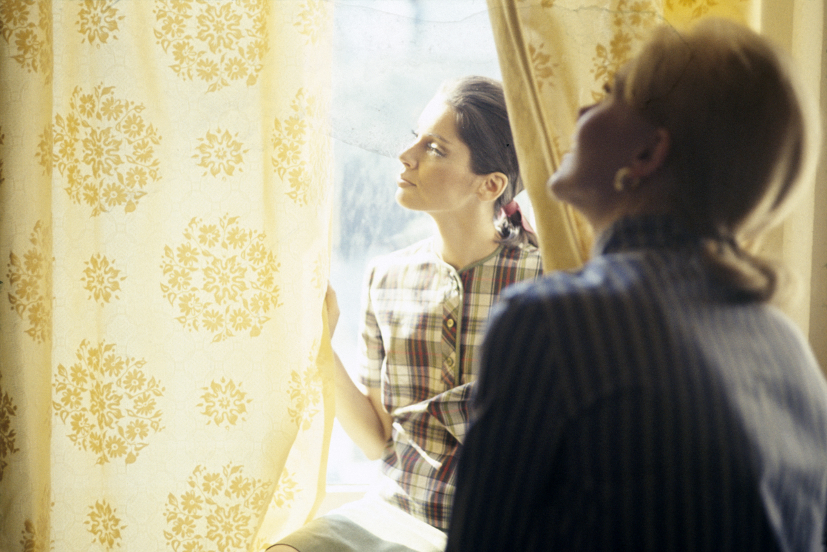 A pair of unidentified models, one in a yellow, red, and black plaid top, look at yellow print curtains in a window, 1965