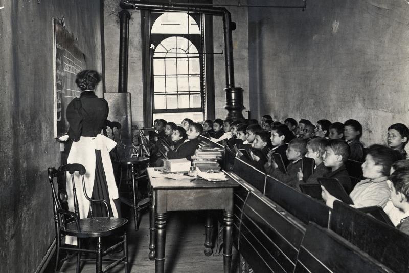 A teacher demonstrates on the blackboard, as students watch attentively from crowded pews, circa 1886
