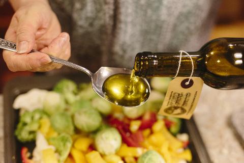Woman pours out olive oil infused with a butter flavoring over a pan of cauliflower, brussels sprouts, red and yellow peppers and broccoli.