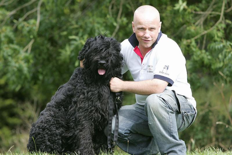 A Black Russian Terrier sits in the grass next to its owner.