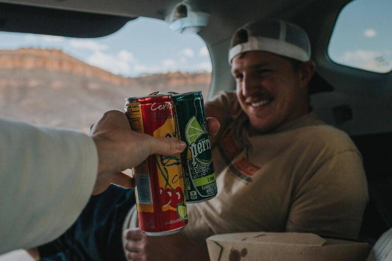 A man hands another man two canned drinks.