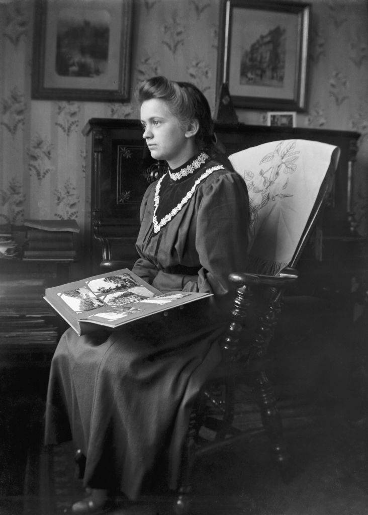 Young girl sitting on ornate wooden chair in living room holding a postcard album circa 1910