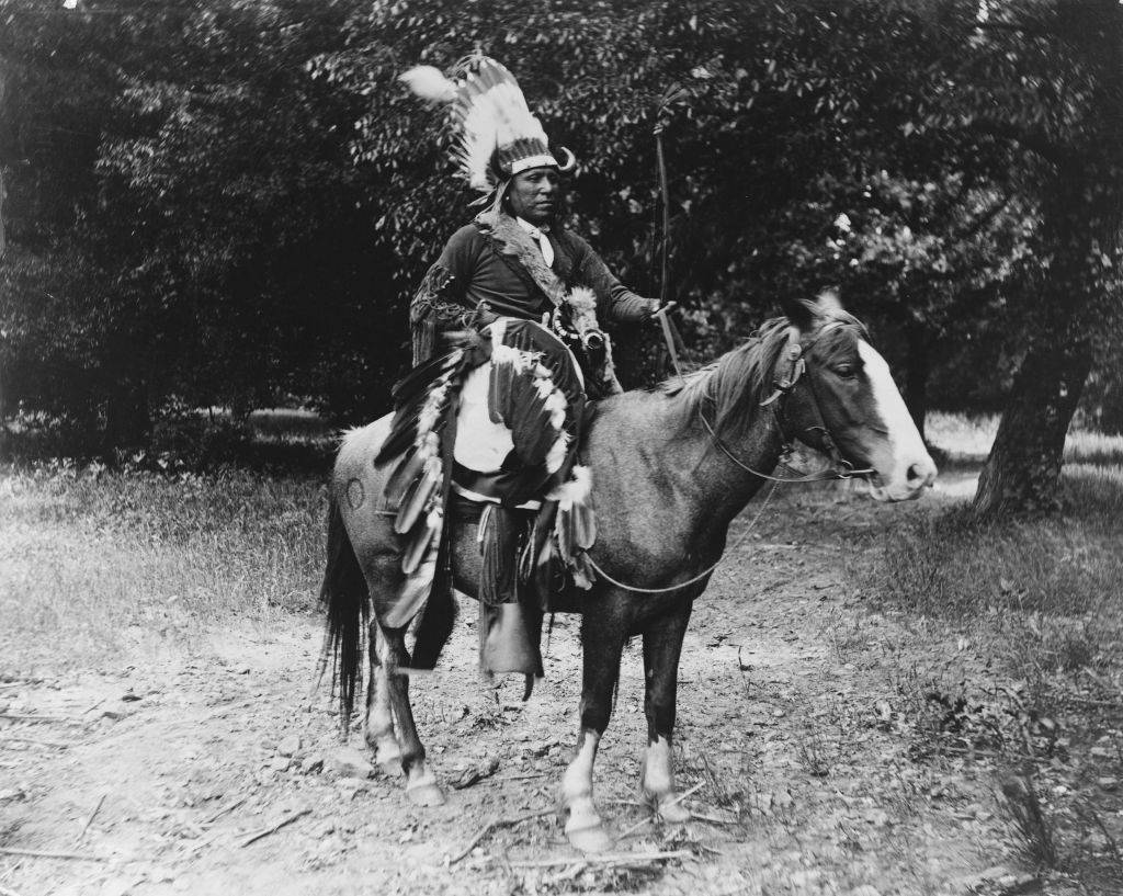 Kiowa warrior Elk Tongue on horseback, wearing a war bonnet, and carrying a shield and bow