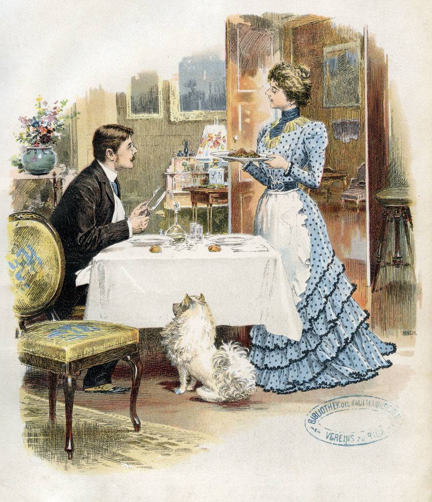 a woman bringing a plate of food to a man who sits at a table with a dog begging at his feet