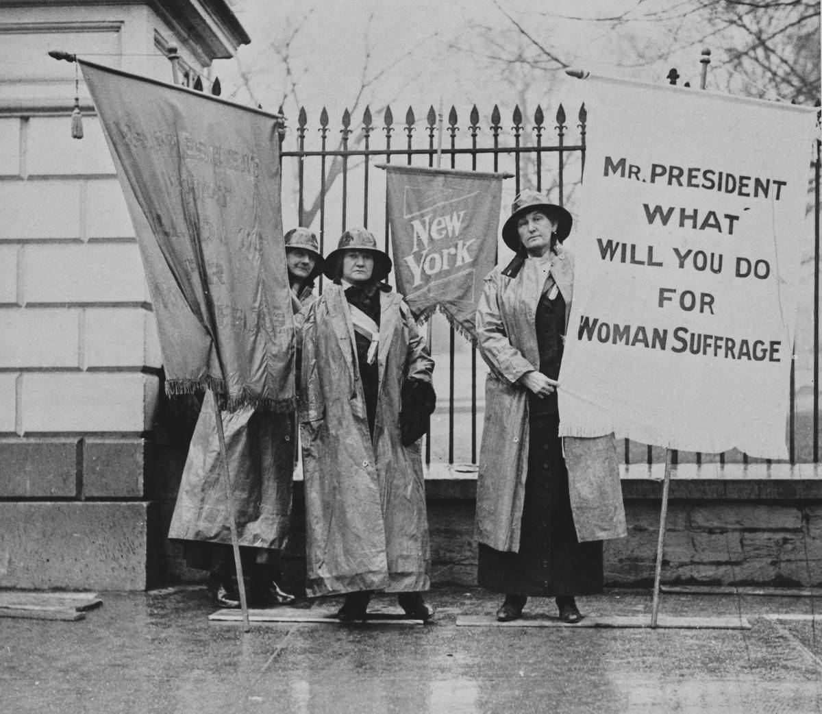 Three women's suffragists protest outside the White House holding a sign that says 