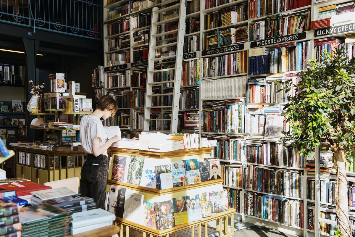 Person looking through book in library piled high with books behind a moving ladder