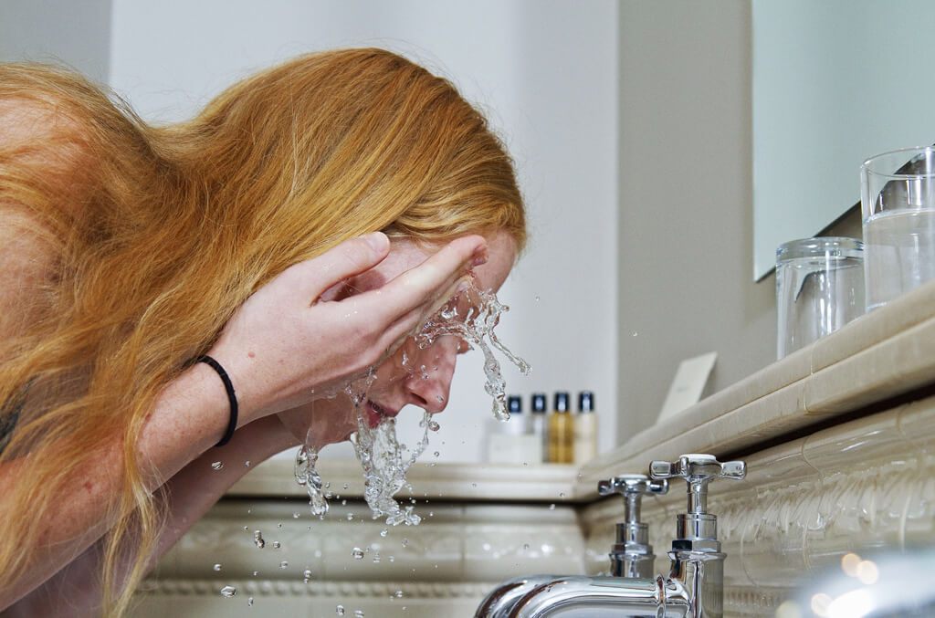 Woman with red hair washing her face over the sink 