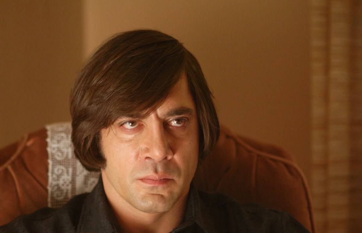 Javier Bardem sitting with angry face as Anton Chigurgh in No Country For Old Men