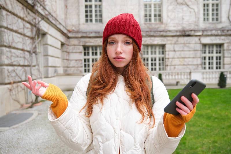 woman shrugging shoulders outside in front of old building while holding phone