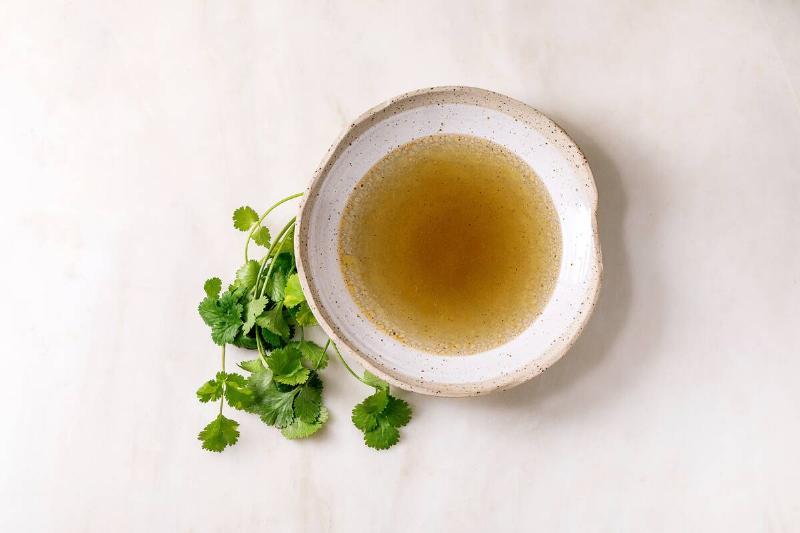 Clear homemade chicken or duck broth bouillon in ceramic bowl with fresh green coriander bunch on white marble table. Flat lay, copy space
