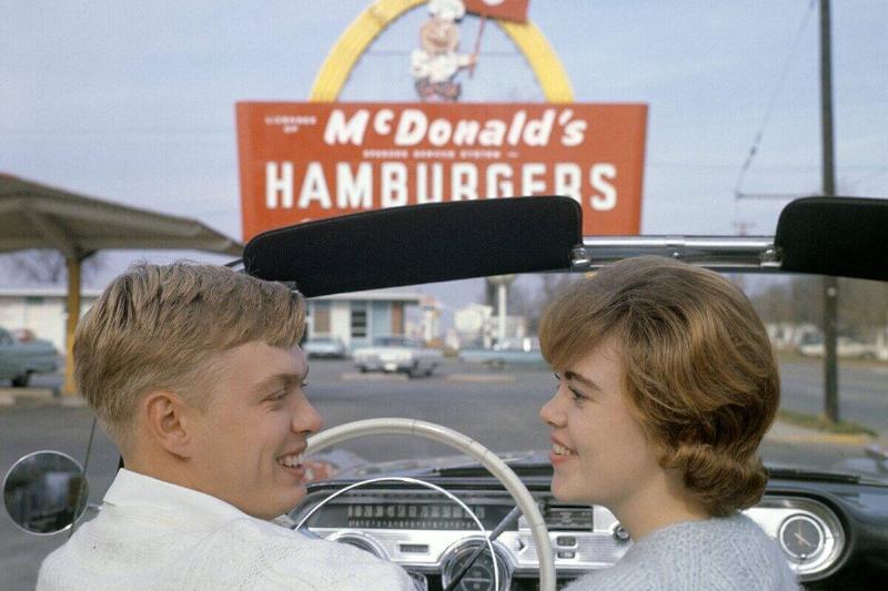 Fast food in the 1950s. A young couple in their convertible car with a McDonald's sign in the backgrou...