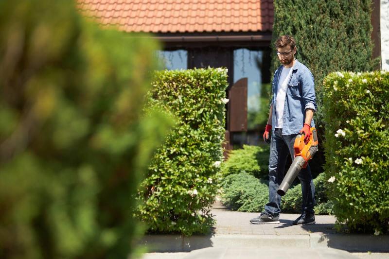 A man in casual outfit removing dry leaves from sidewalk with hand blower in sunny day.