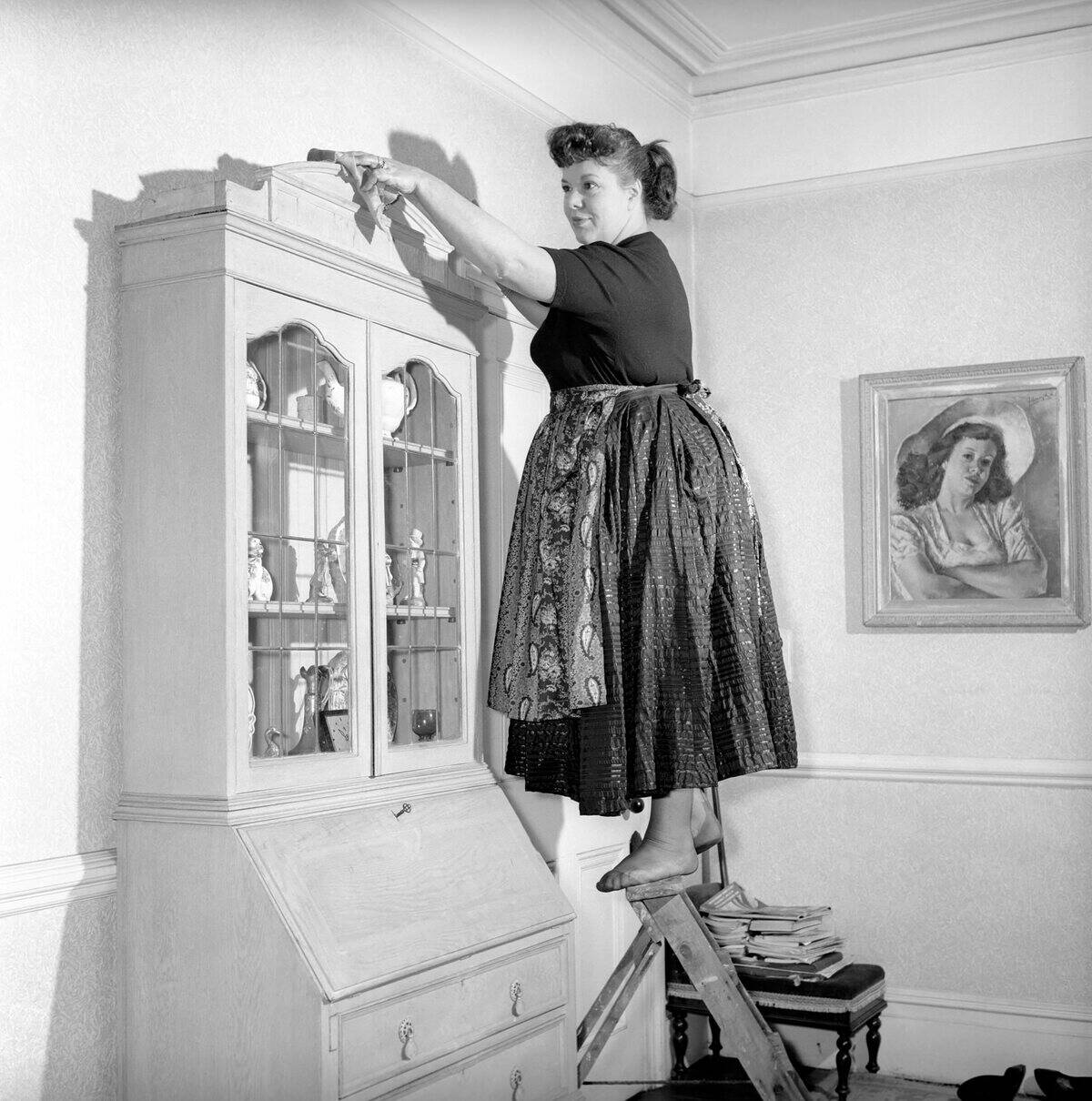 Mrs. Olive Haugh dusting the bookcase as she completes the daily household chores, circa 1954