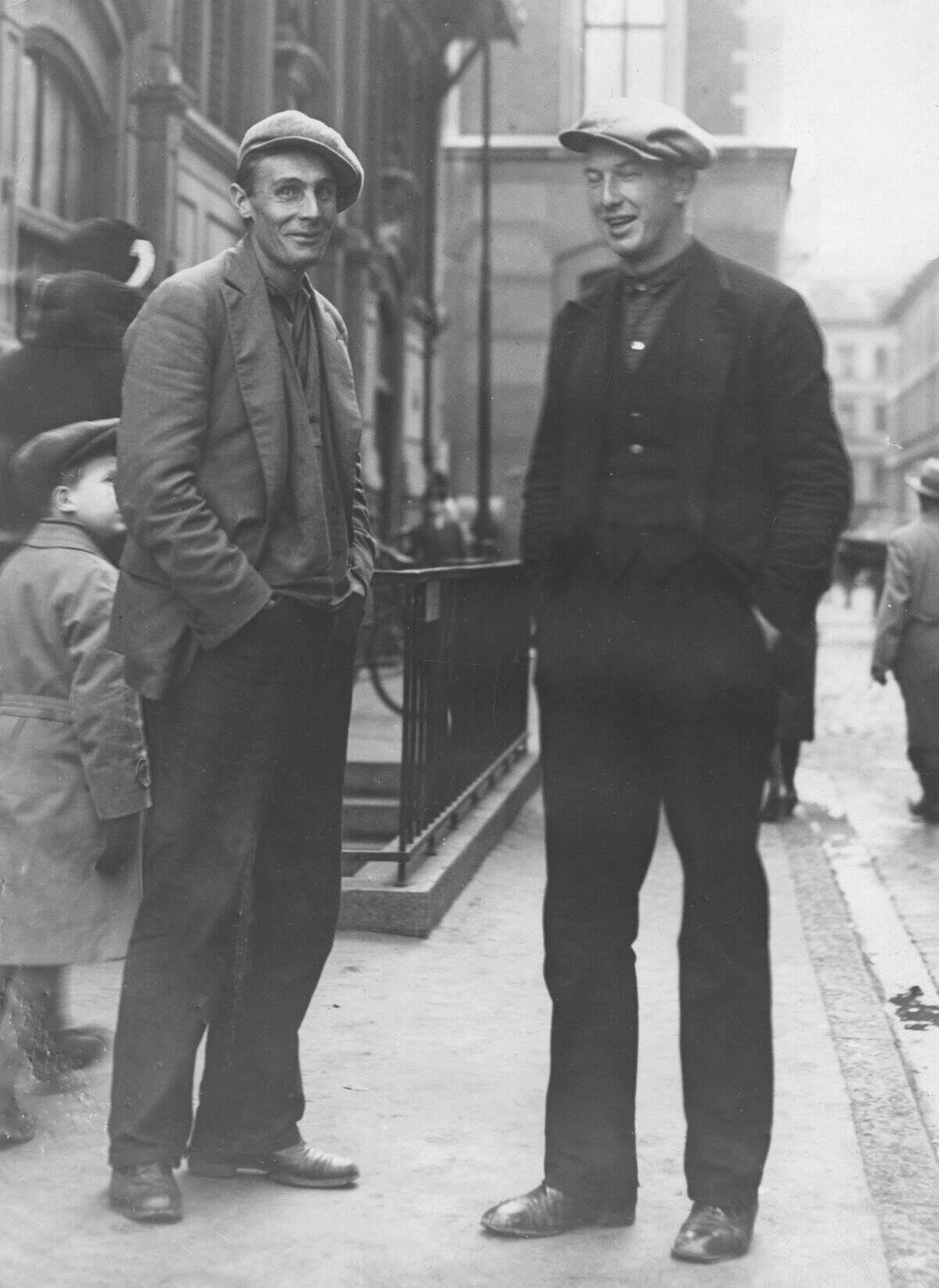 Two simply dressed workers stand on a street in Stockholm with their hands in their trouser pockets. I...