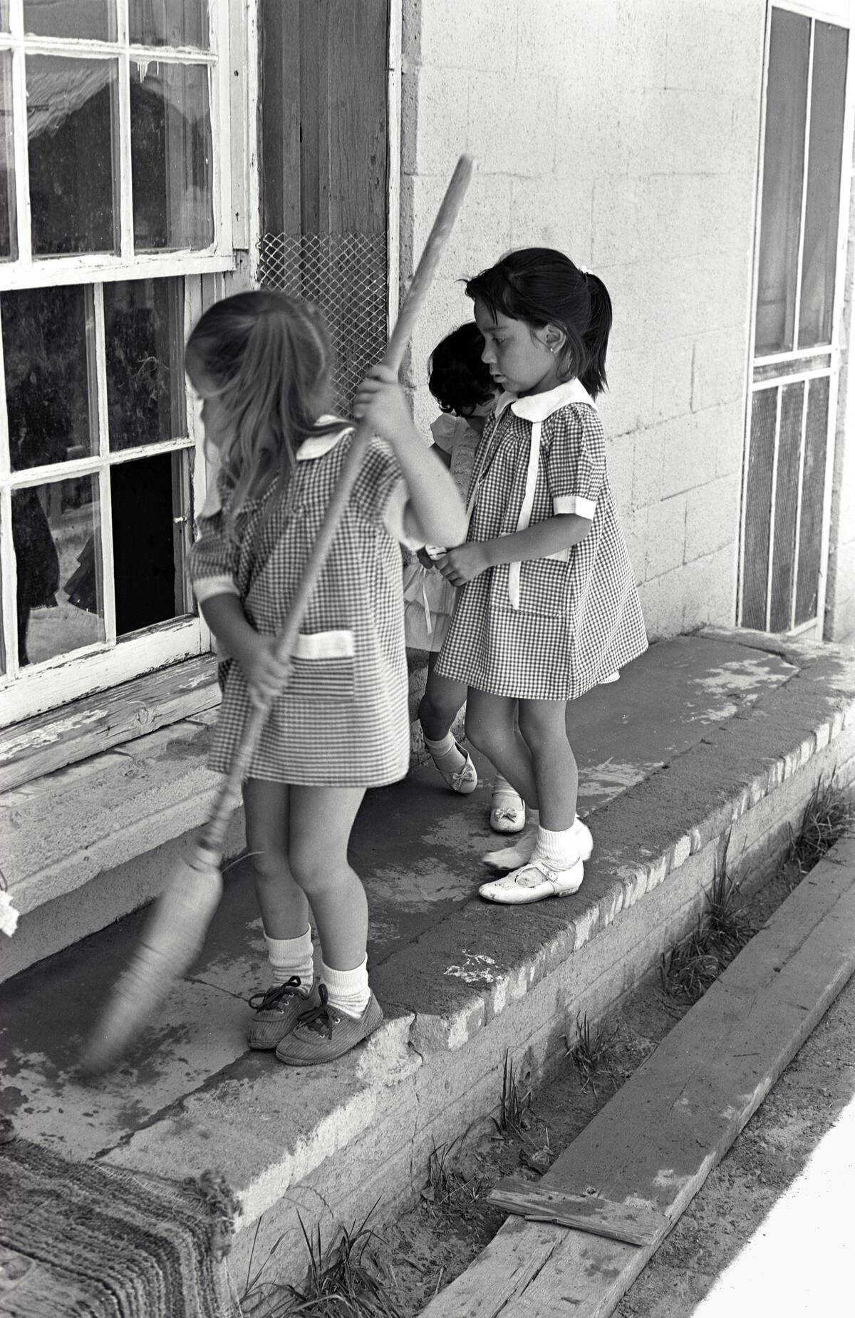 Two Young Girls Sweep A Porch