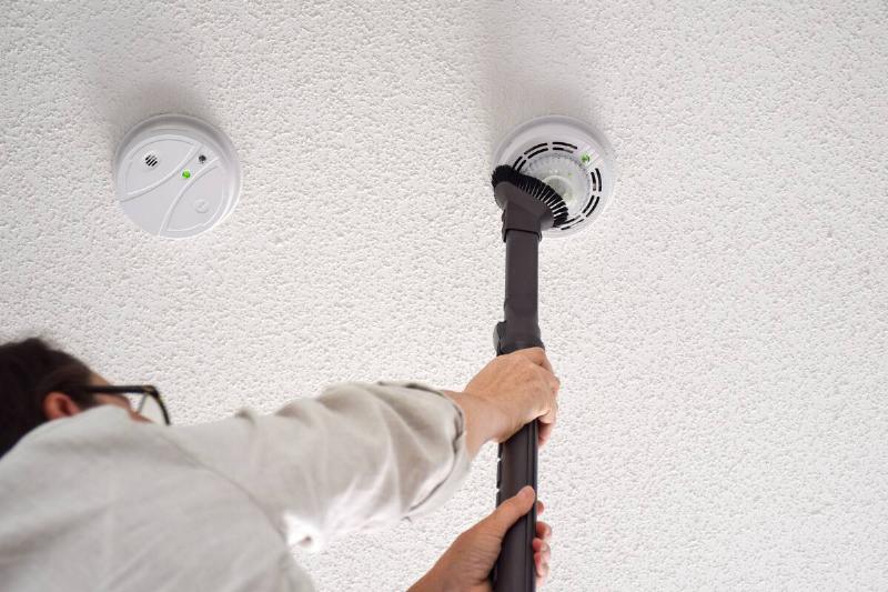 Woman vacuuming smoke and carbon monoxide detectors on a ceiling of a domestic room.