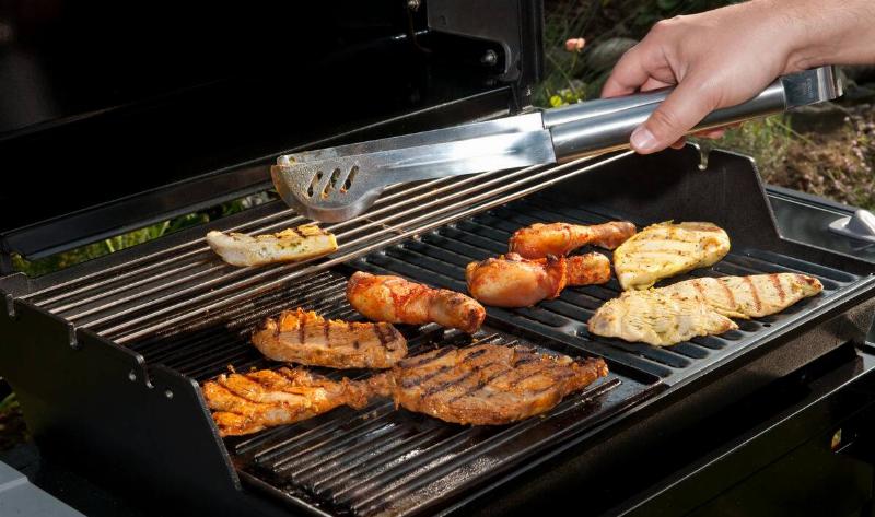 A barbeque grilling a variety of meats, a hand reaching in with some metal tongs.