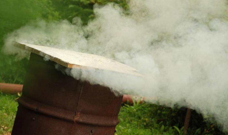 A burn barrel with a piece of wood over it, smoke pouring out of it.