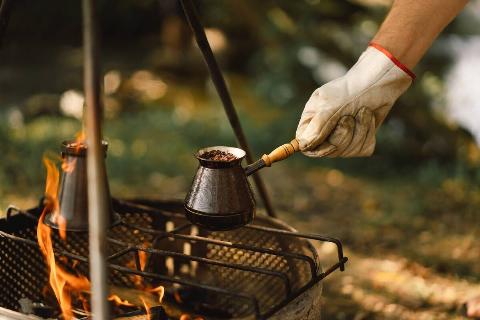 A gloved hand making coffee over a fire outdoors.