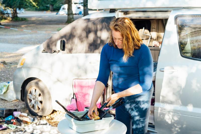 Middle age nomad woman washes dishes outside her camper van in forest camp during road trip.
