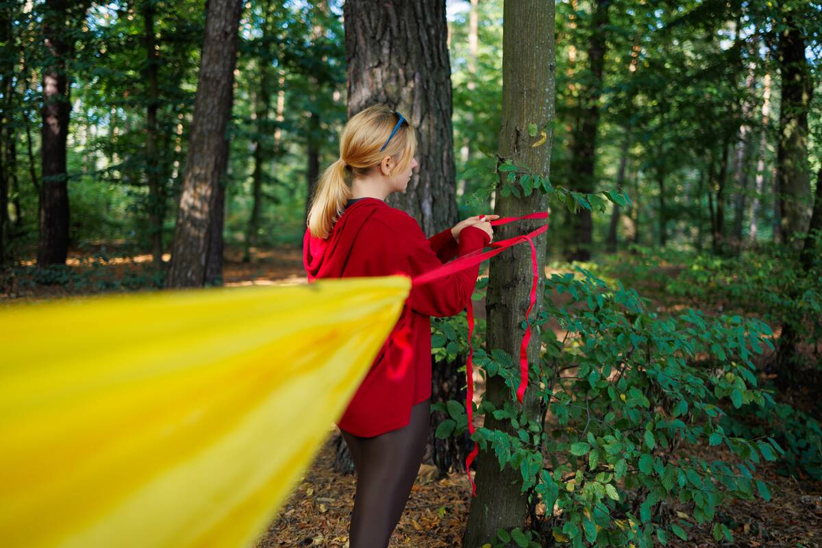 A red-haired woman in a red hoodie, leggings and glasses on her head hooks a hammock to the trees in the forest
