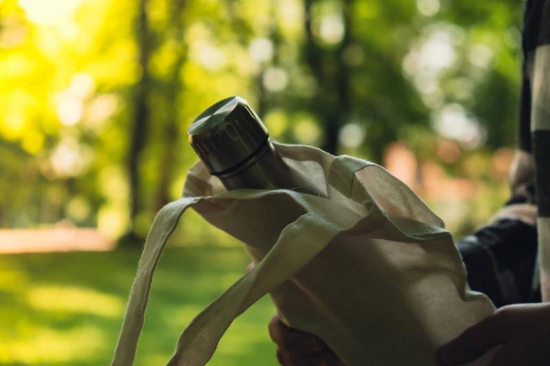 Close-up of Unrecognizable female hand holding Water bottle from eco bag. Reusable steel thermo water bottle in park.