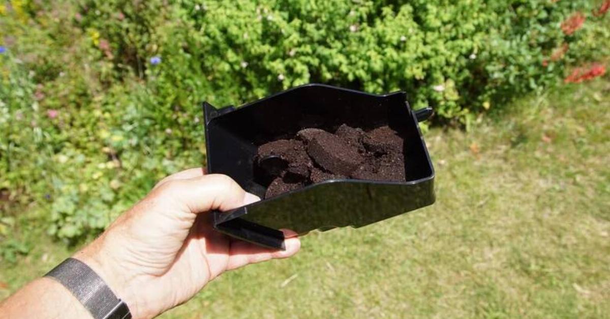 A hand holding a container of coffee pucks near a garden.