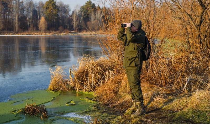 A hunter standing on the water's edge, looking through some binoculars.