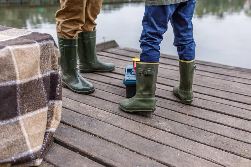 Father and son in rubber boots standing on wooden pier at lake.