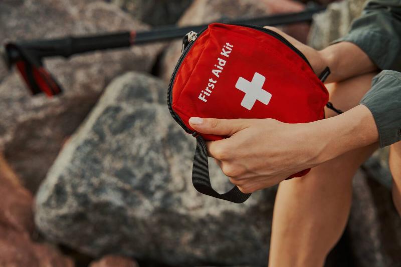 Hiker holding a red medical kit with both hands while resting on rocks near the shore