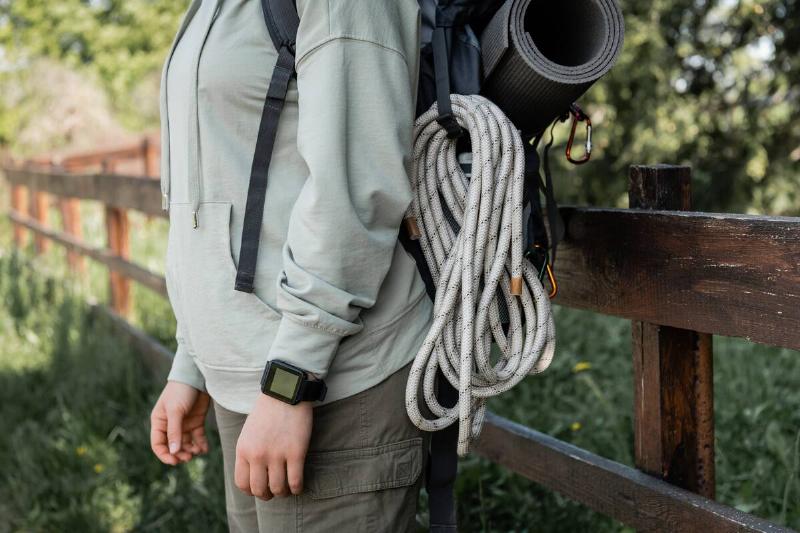 Cropped view of young female hiker with fitness tracker, backpack and travel equipment standing next to a wooden fence, a roll of rope on her side.