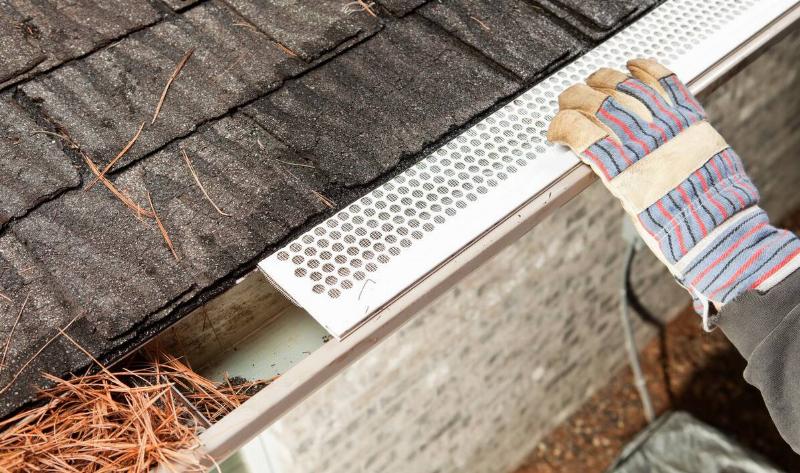 A gloved hand installing a gutter guard on gutters filled with pine needles.