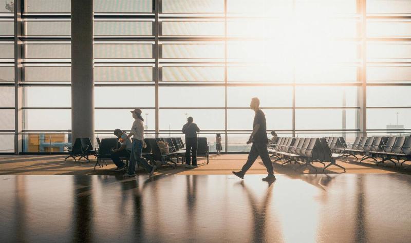 Some people walking through a large, open airport terminal as the sun rises through the window.