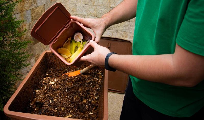 A person emptying a small container of food scraps into a fresh compost bin.