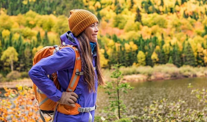 A woman hiking in fall weather, smiling, with a coat, beanie, and gloves on.