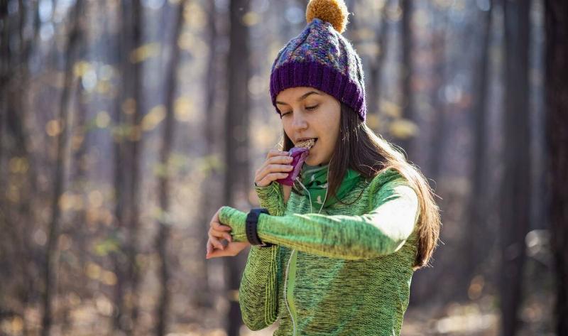 A girl standing in a forest, eating a granola bar and checking her watch.