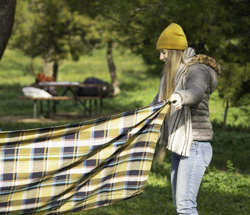 Smiling young woman preparing picnic area in a park, holding a checkered blanket