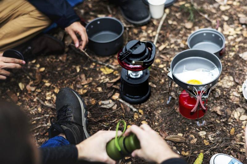 A small camp stove and pan on the ground next to a lantern, people sitting around it, an egg in the pan.