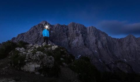 A person hiking at night with a headlamp, standing on a rocky outcrop.