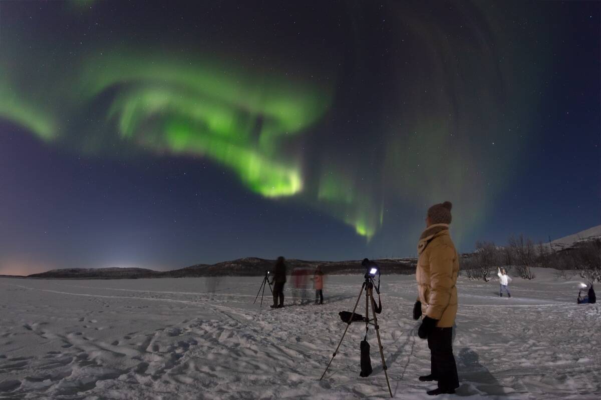 A group of people in a snowy field with camera setups, taking photos of the northern lights.
