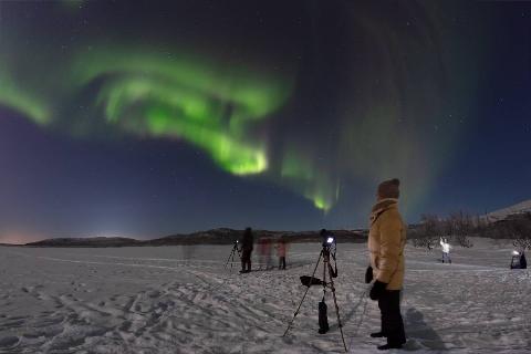 A group of people in a snowy field with camera setups, taking photos of the northern lights.