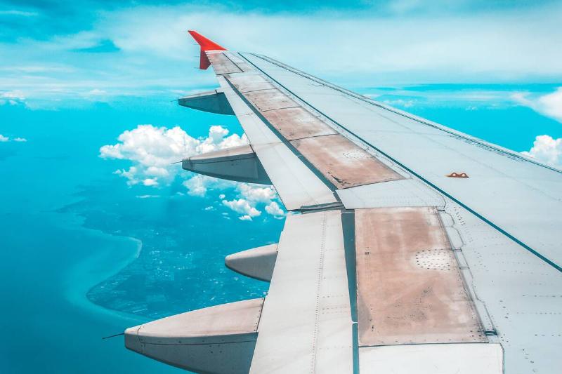 The wing of a plane as it flies over the ocean, a yellow hook latch visible.