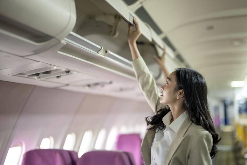 A woman is looking into the overhead compartment of an airplane. She is wearing a suit and she is in a hurry