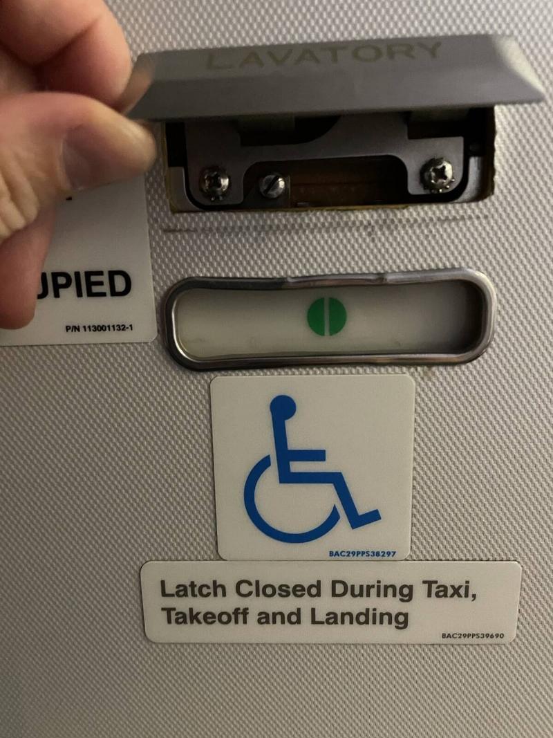 A close photo of a plane's bathroom door signage, a hand lifting up the 'lavatory' sign to show a secret latch.
