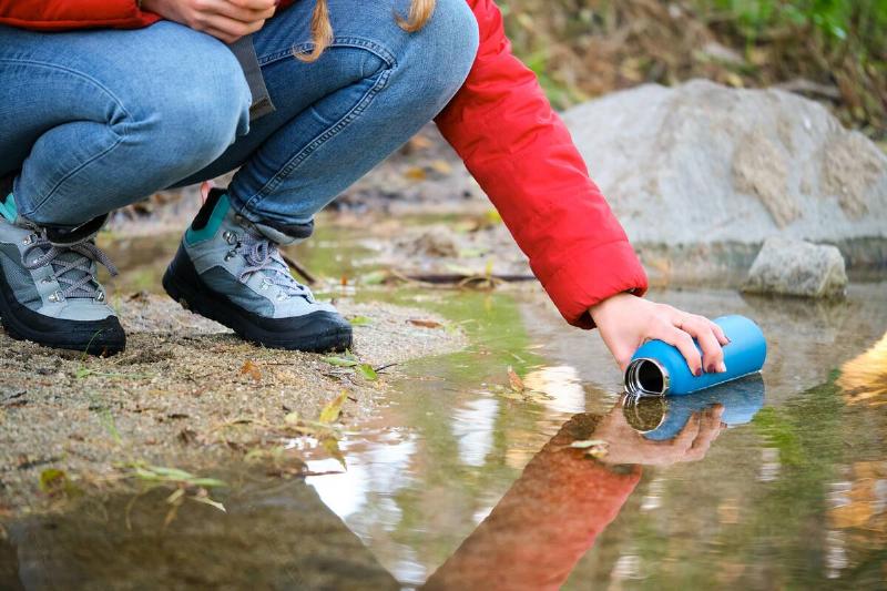 Unrecognizable female trekker filling water bottle of raw water in a river.