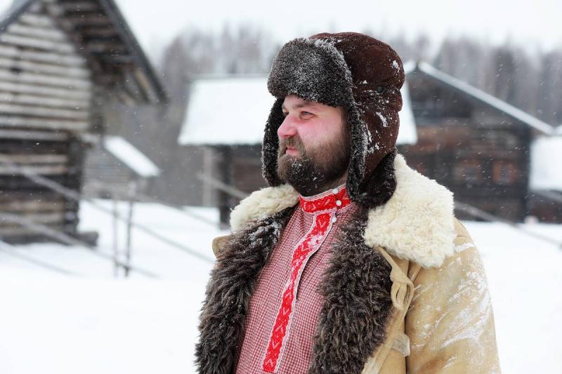A bearded man standing in the snow with a trapper style hat on, as well as a thick jacket.